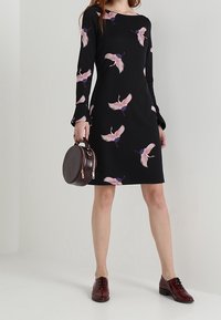 Robe noire à manches longues, ornée de motifs de grues rose et violet. Associée à des chaussures bordeaux et un sac rond. Texture lisse.
