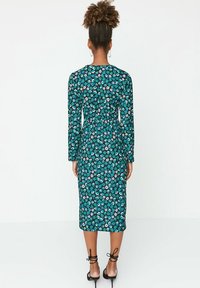 Femme aux cheveux bouclés en chignon haut portant une robe midi à manches longues florale teal et noire, ainsi que des sandales à talons hauts noires, se tenant devant un mur blanc.