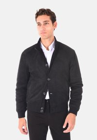 Giacca bomber nera in suede con colletto alto, chiusura a bottoni, polsini a coste e design aderente. Indossata sopra una camicia bianca.