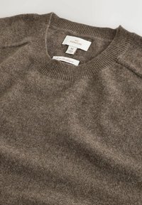 Brauner Wollpullover mit Rundhalsausschnitt und gerippten Bündchen. Die Stofftextur wirkt weich und gestrickt. Innen sind Etiketten sichtbar, die Marke und Größe anzeigen.