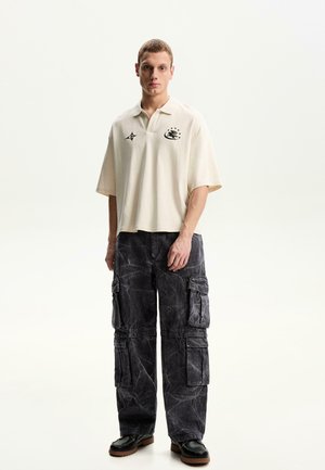 Tricou polo crem cu mâneci scurte, având accente grafice negre. Pantaloni cargo negri cu un finisaj texturat și multiple buzunare. Pantofi negri.