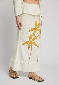White cotton skirt with yellow embroidered palm leaves, frayed hem, and a tie waist. Model wears beige sandals.