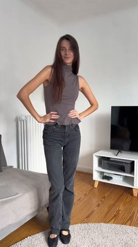 Grijze mouwloze top met een schijnbare hals; zwarte rechte jeans; zwarte schoenen; staande pose; lichte achtergrond met meubels.