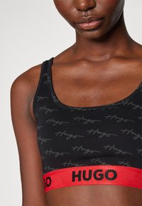 Melna sporta krūštura iezīmē atkārtojošu "Hugo" logotipa rakstu, sarkano elastīgo jostu ar "HUGO" tekstu un apaļu dekoltē. Mīksta auduma struktūra.