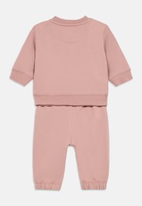 Rozā divdaļīgā sporta tērpa komplekts, kas izgatavots no mīkstas kokvilnas. Raksturo apaļš dekoltē, garas piedurknes, ribotas aproces un elastīga jostasvieta ar sašaurinātām kājām.