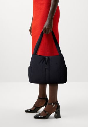 Bolso de abrigo en color marino con dos asas, que cuenta con un cierre de cremallera y bolsillos exteriores. El modelo lleva un vestido rojo y tacones de charol negros.