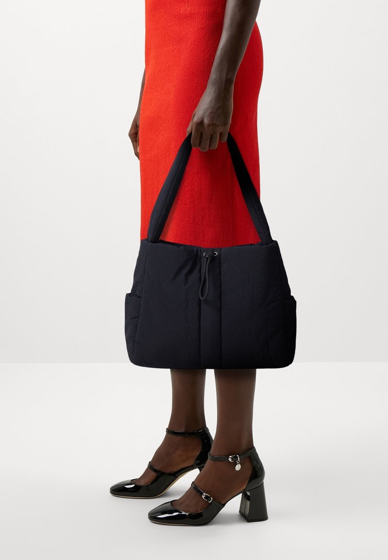 Sac à main rembourré bleu marine avec deux poignées, doté d'une fermeture éclair et de poches extérieures. Le modèle porte une robe rouge et des escarpins noirs en vernis.