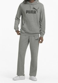 Sudadera de algodón gris con un bolsillo delantero y cordón, que presenta un logo negro de "PUMA". Pantalones de chándal grises a juego con un pequeño logo en el muslo. Zapatillas blancas.