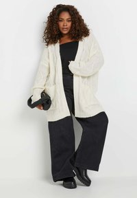 Cremfarbener Strickcardigan mit lockerer Passform, der ein Kabelstrickmuster, Taschen und gerippte Bündchen aufweist, kombiniert mit hoch taillierten schwarzen weit geschnittenen Hosen.