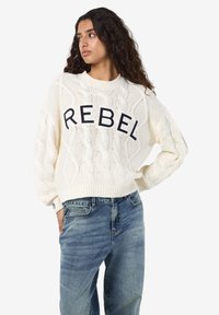 Kreminė kabelinės virvės megzta palaidinė su žodžiu "REBEL" tamsiai mėlyna spalva per visą priekį. Ripiavimo rankogaliai ir apačia. Derinama su džinsiniais džinsais.