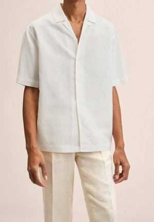 Homme portant une chemise blanche ample à manches courtes boutonnée et un pantalon beige bicolore, debout devant un fond uni.