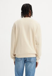 Beige collegegenser med løs passform, lange ermer og ribbede mansjetter. Sammen med lyseblå jeans, som viser et enkelt, uformelt design.