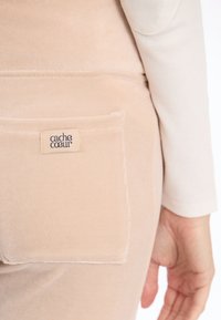 Pantalons en velours beige avec une poche arrière portant une étiquette. Texture lisse et détails de couture soignés. Chemise légère à manches longues visible.