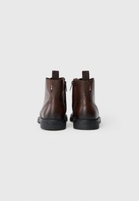 Cizme de gleznă din piele maro cu o textură netedă, prezentând detalii decorative de brogue, închidere cu fermoar și o talpă din cauciuc negru.