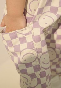 Lila- und cremefarbener karierteter Stoff mit einem Muster aus lächelnden Gesichtern. Eine Hand steckt in einer seitlichen Tasche und zeigt die Struktur der Tasche.