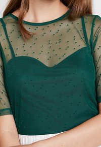 Blusa verde oscuro con una superposición transparente de puntos. Presenta un escote en forma de corazón y mangas cortas, con textura y patrón contrastantes.