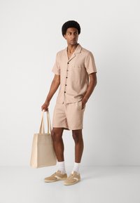 Camisa de manga corta de color marrón claro con un patrón sutil, combinada con pantalones cortos a juego, bolsa de tela beige y zapatillas de color tostado. Tela suave y diseño informal.