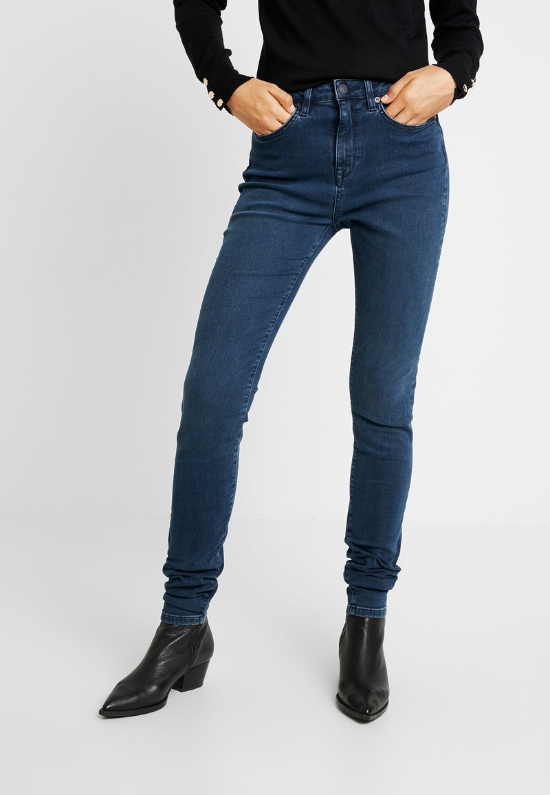 Jean skinny en denim bleu foncé, taille haute, avec un style classique à cinq poches et une légère décoloration. Associé à des bottines noires.