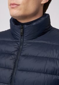 Marineblauer Steppjacke mit hohem Kragen, gestepptem Design, glatter Textur und einem Frontreißverschluss. Rippstrick-Detail am inneren Kragen.