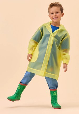 Niño sonriente con un impermeable amarillo translúcido, jeans azules y botas de goma verdes con borde rojo, posando frente a un fondo beige.