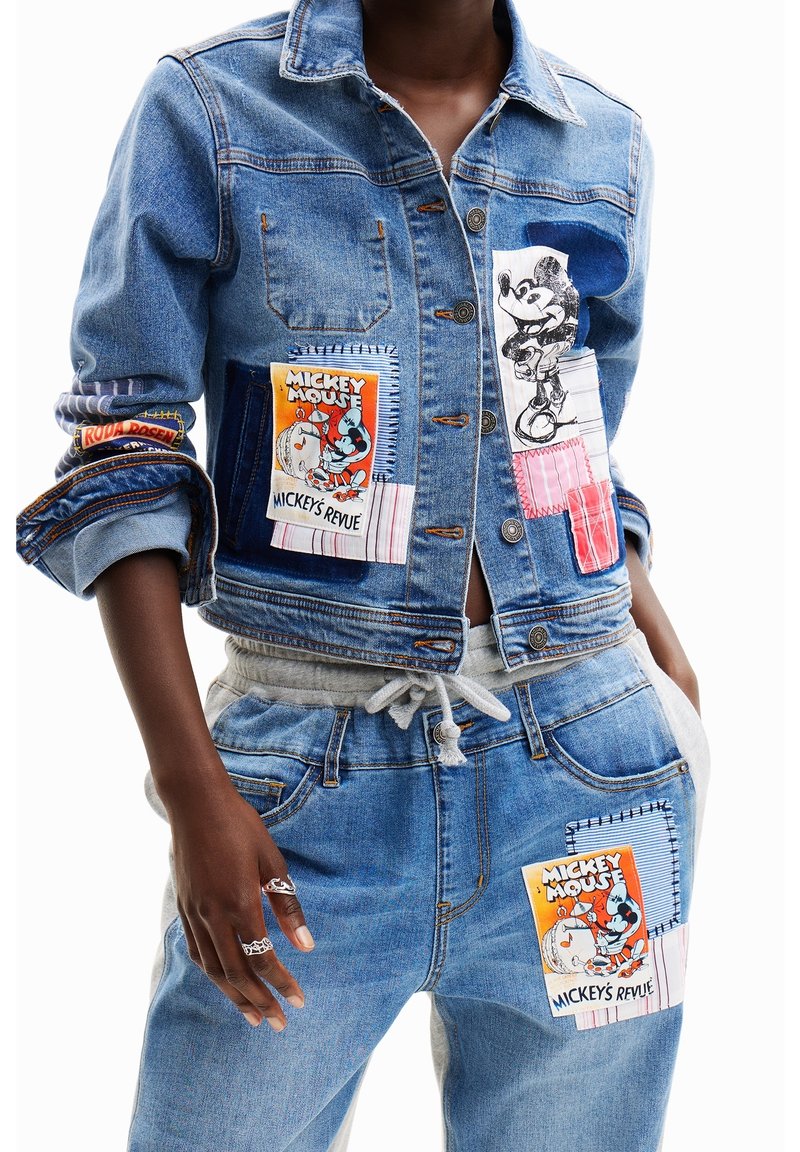 Jeansjacka med lappar som visar Mickey Mouse-grafik; blått tyg, knäppning framtill och olika texturer inklusive sydda detaljer på ärmarna.