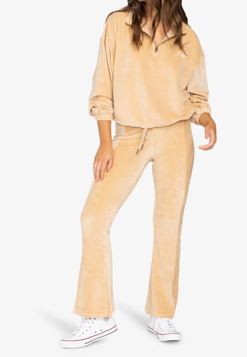 Personne portant un pantalon évasé en velours côtelé beige et un pull assorti à demi-fermeture éclair, associé à des baskets montantes blanches sur fond blanc.