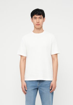 Joven de pie frente a un fondo blanco, vistiendo una camiseta blanca de manga corta lisa y pantalones vaqueros azul claro, mirando hacia adelante con expresión neutral.