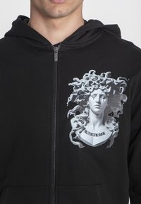 Crna zip-up dukserica s kapuljačom koja prikazuje grafiku Meduze s detaljnom kosom, teksturiranim materijalom i prednjim džepom. Dizajn uključuje riječ "Kanalag."