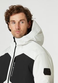Vitt och svart vattentät jacka med hög krage, blixtlåsstängning, kantig design och märke på ärmen; har ett slätt, strukturerat tyg.