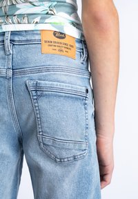 Jeans de mezclilla azul claro con bolsillo parche, detalles de costura y etiqueta de cuero marrón. La tela tiene una textura desgastada y un aspecto envejecido.