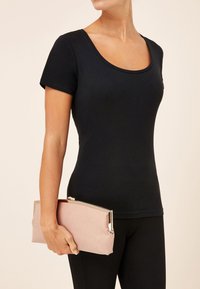 Femme portant un haut noir ajusté à manches courtes et un pantalon noir, tenant une pochette rectangulaire rose poudré avec un cadre métallique.