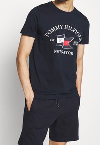 Navy blue cotton T-shirt featuring a graphic logo with flags and text. Crew neck, short sleeves, relaxed fit. Paired with navy shorts.