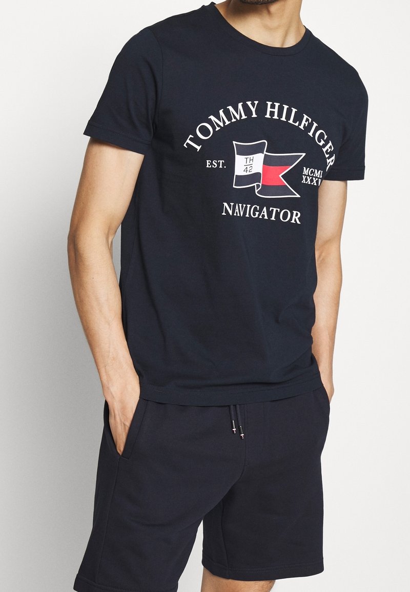 Navy blue cotton T-shirt featuring a graphic logo with flags and text. Crew neck, short sleeves, relaxed fit. Paired with navy shorts.