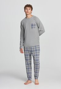 Maglietta da pigiama grigia a maniche lunghe con taschino a quadri, abbinata a pantaloni grigi a quadri con motivi a righe blu e neri. Materiale in cotone.