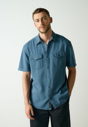 Homme aux cheveux courts et à la barbe portant une chemise bleue à manches courtes avec deux poches poitrine, debout devant un fond clair uni.