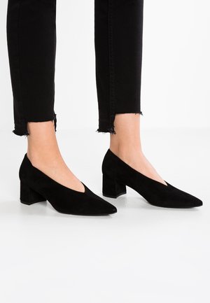 Sorte ruskind sko med lav blokhæl båret med sorte cropped skinny jeans med frynset kant på hvid baggrund.