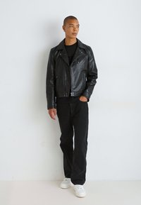 Must mustrõngast mehhanismiga must nahast mootorrattakuub, millel on ebaühtlane lukk, nööpkaelus ja kaks esikotti, kombineeritud mustade teksade ja valgete tennisetossudega.