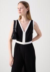 Combinaison noire avec décolleté en V et bordure blanche. Tissu plissé de la taille à l'ourlet. Portée avec un petit sac à main noir. Texture lisse.