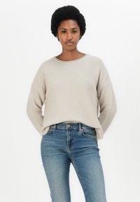 Sweater de malha bege claro com corte solto, decote arredondado e costuras de ombro caídas, combinado com jeans skinny azuis. Design simples, textura suave.