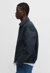 Giacca da bombardiere blu navy con colletto, polsini in elastico e vita elasticizzata. Presenta una toppa con logo sulla manica sinistra. Texture liscia.