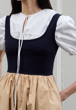 Woman wearing a layered dress with a white puff-sleeve top, black sleeveless bodice, and beige gathered skirt, standing with hands on hips.