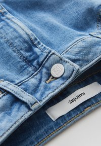 Lichtblauwe spijkerbroek met zilveren knoopsluiting, zichtbare naden en een stoffen label met de merknaam.