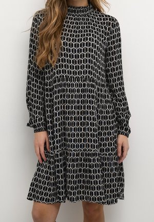 Femme portant une robe à manches longues à motif géométrique noir et blanc avec un col montant et une jupe à volants.