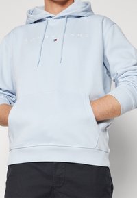 Světle modrá mikina s kapucí z jemné látky, vybavená přední kapsou, žebrovanými manžetami a bílým logem "TOMMY JEANS" přes hrudník.