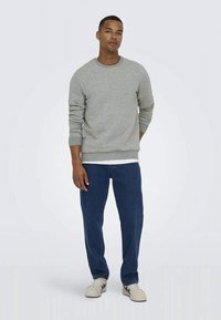 Jovem de pé, vestindo um moletom cinza, jeans azuis e ténis brancos, em frente a um fundo branco simples.