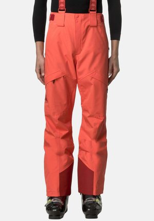 Pantaloni da sci rosso corallo brillante con bretelle, tasche con zip sulle cosce e rinforzi interni di colore rosso scuro nella parte inferiore delle gambe, indossati con scarponi da sci neri.