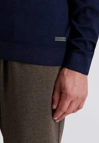 Donkerblauwe gebreide trui met een geribde zoom, gecombineerd met gestructureerde bruine broeken; met een klein grijs merklabel op de trui.