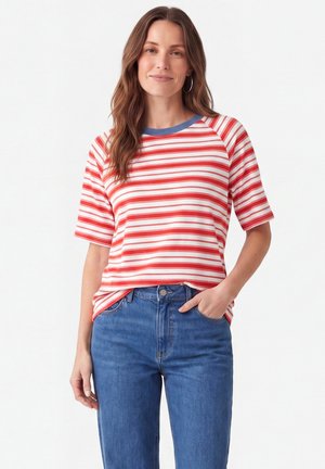 Kvinde med langt brunt hår iført en rød og hvid stribet kortærmet trøje og blå jeans, der står med den ene hånd i lommen mod hvid baggrund.