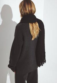 Czarny oversize sweter z prążkowanym dołem i mankietami, w połączeniu z dopasowanym szalikiem z frędzlami. Miękka, teksturowana tkanina o przytulnym wyglądzie.