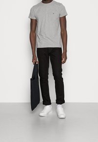 T-shirt grigia a maniche corte con un piccolo logo, abbinata a jeans neri slim-fit e sneaker bianche. In mano tiene una borsa tote nera.